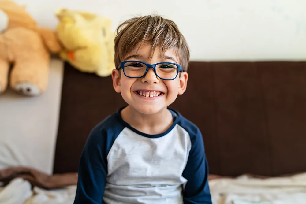 a little boy sitting on his bed smiling and wearing glasses