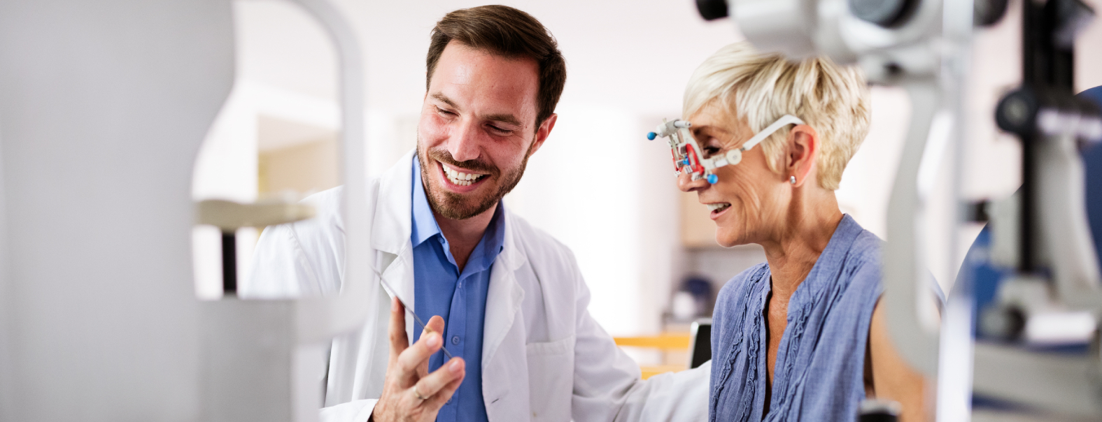Banner Image Home An eye doctor smiles while explaining results to a patient wearing diagnostic eye testing equipment during an eye exam.