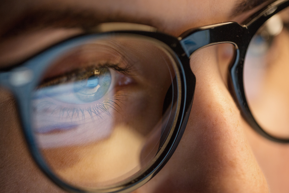 Close-up of a person wearing glasses, reflecting a computer screen on the lenses, with eyes focused intently on the display.