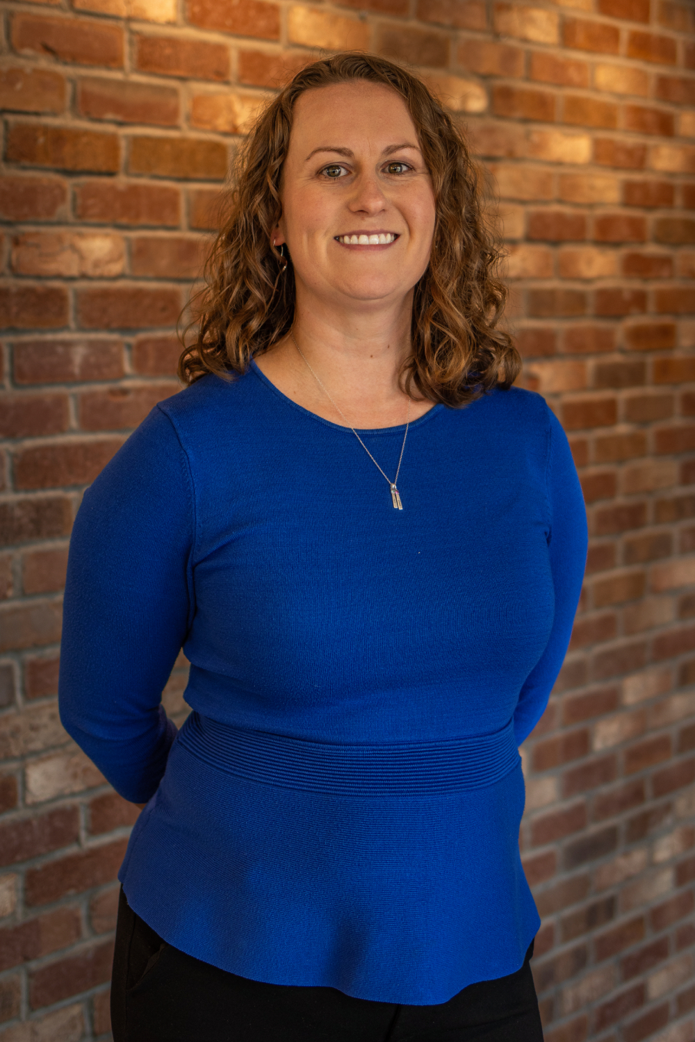 Caitlin A woman with curly hair wearing a blue long-sleeve top stands in front of a brick wall, smiling at the camera.