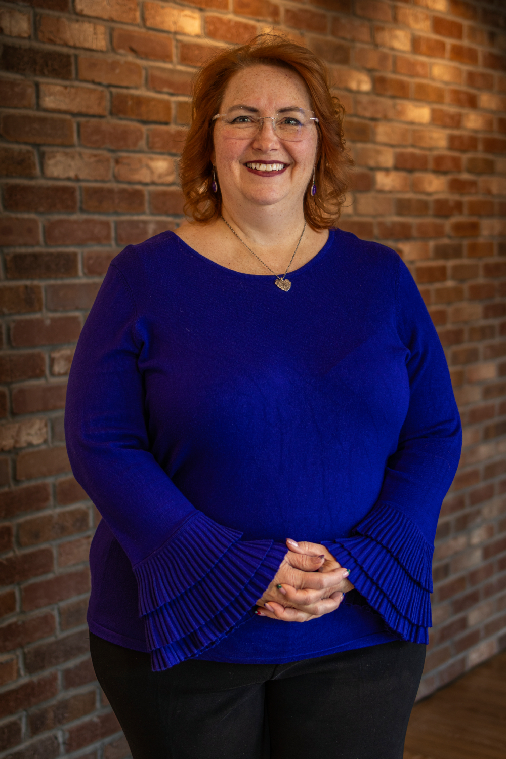 Char A woman with red hair, wearing glasses and a blue blouse with pleated sleeves, stands in front of a brick wall, smiling with her hands clasped.