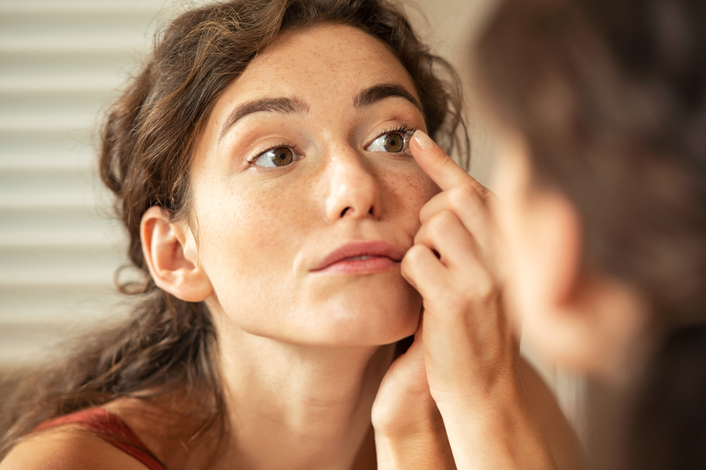 A woman looks in a mirror and places a contact lens onto her right eye using her finger.