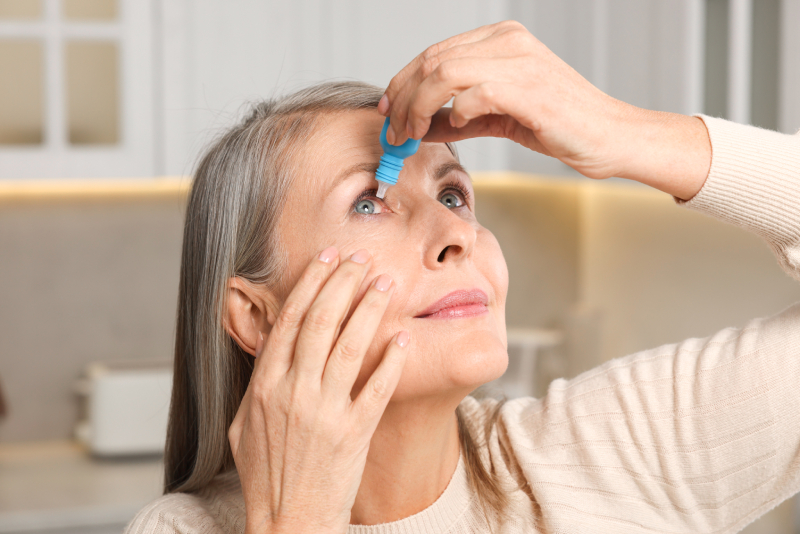 Dry Eye Treatment Older woman with gray hair applies eye drops to her right eye in a modern kitchen setting.