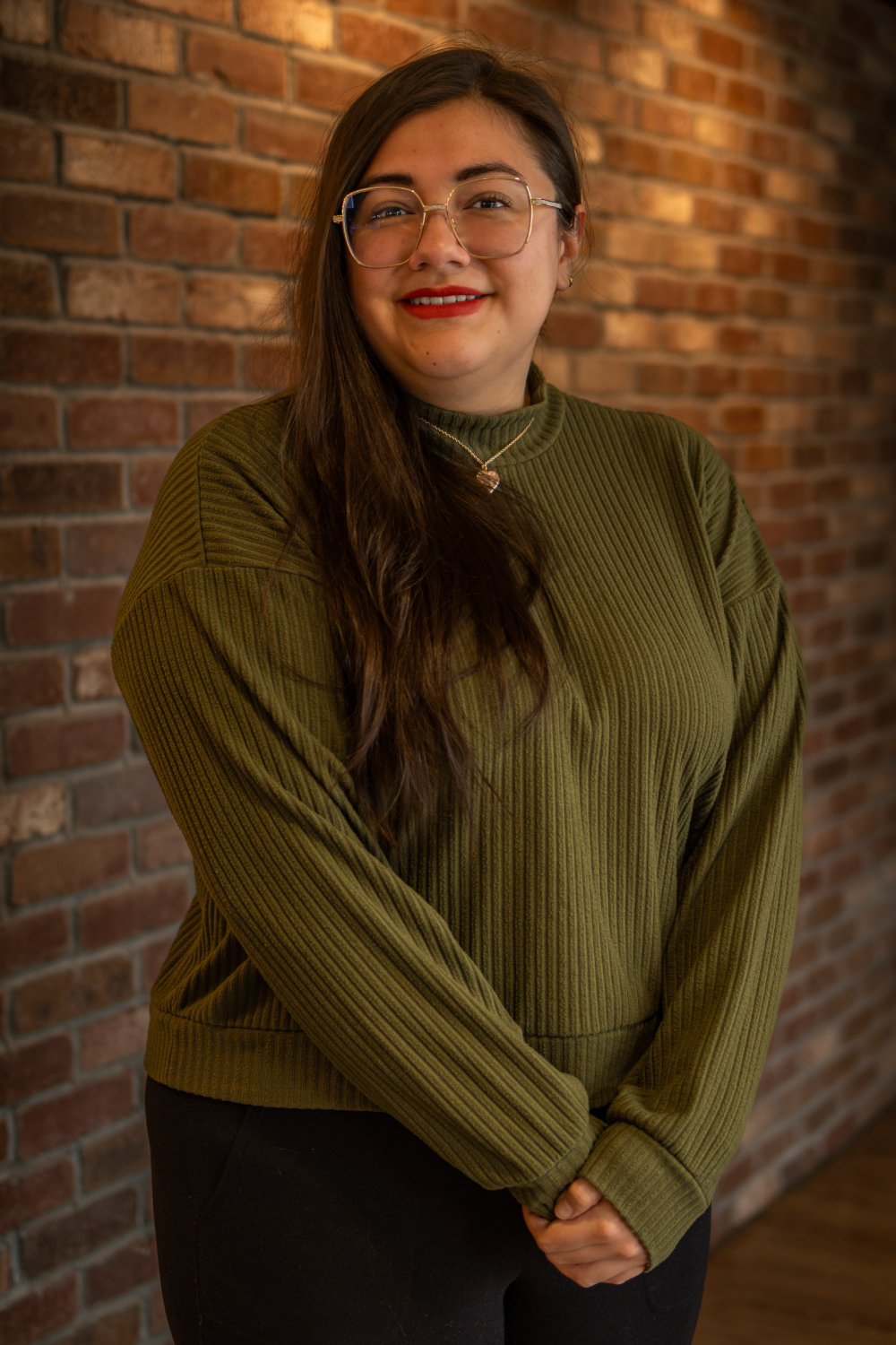 Karlie A person with long brown hair, wearing glasses and a green ribbed sweater, stands in front of a brick wall, smiling slightly with hands clasped.