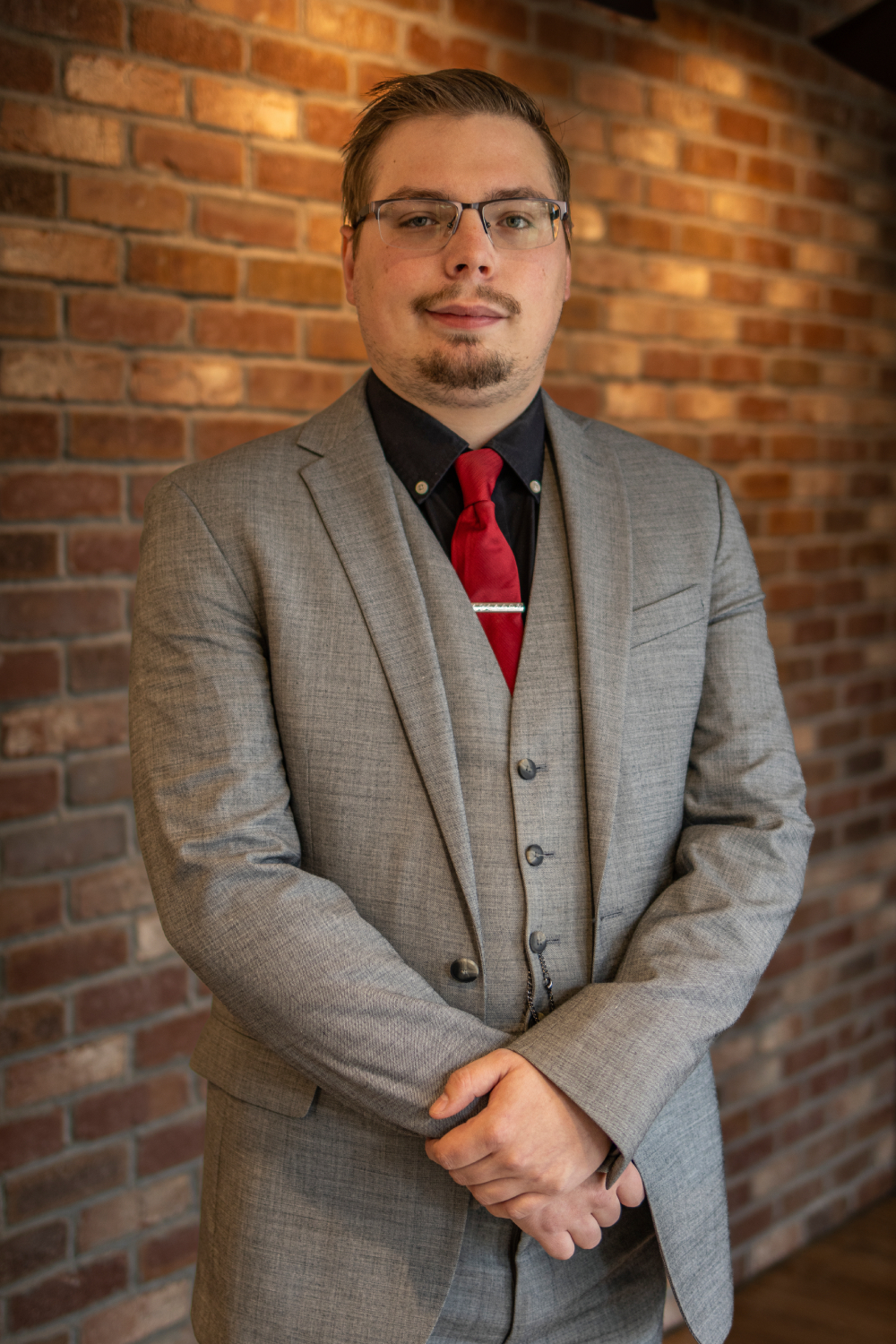 Kellen A man in a gray suit, black shirt, and red tie stands in front of a brick wall with his hands clasped.