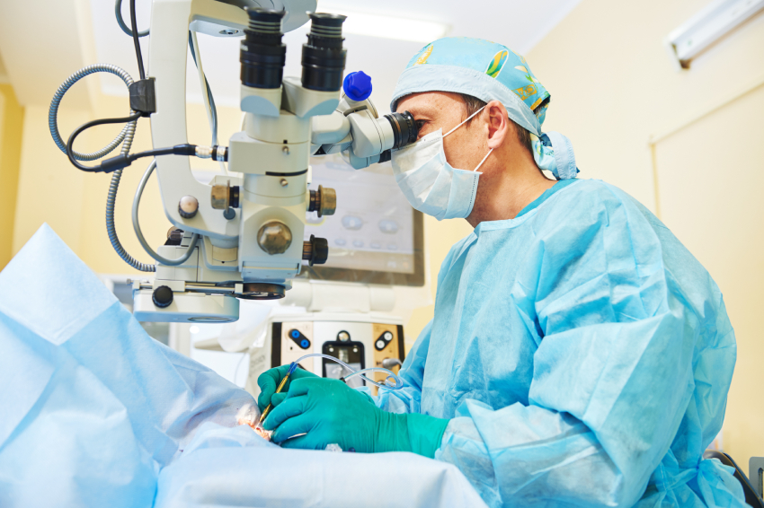 A surgeon in scrubs and mask performs a procedure using a microscope in an operating room.