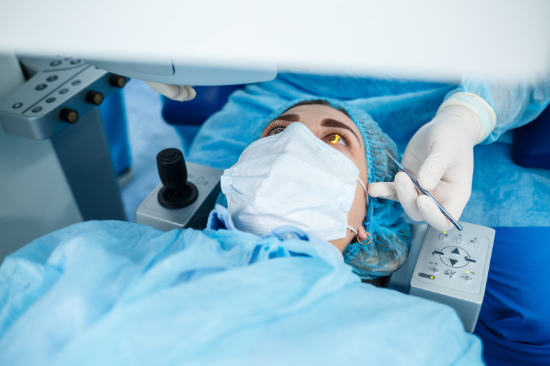 LASIK Consultations A patient wearing a surgical cap and mask lies under a medical machine while a doctor performs an eye procedure using surgical instruments.