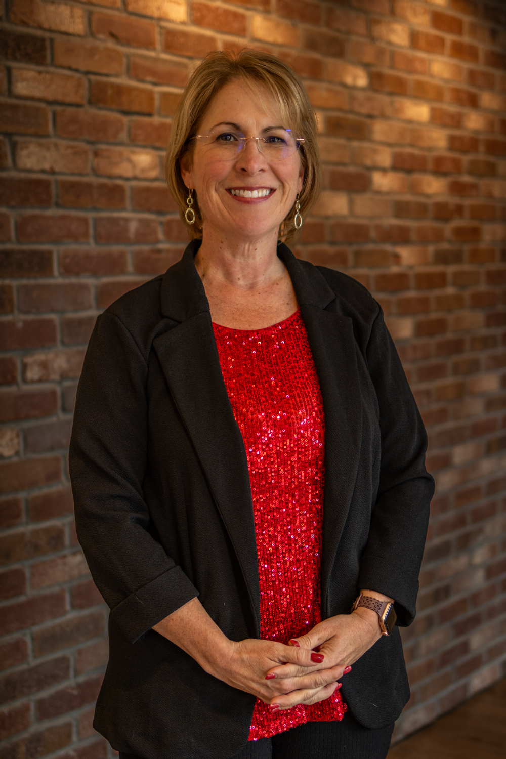 Lynn A woman wearing glasses, a black blazer, and a red sequin top stands in front of a brick wall, smiling with her hands clasped.