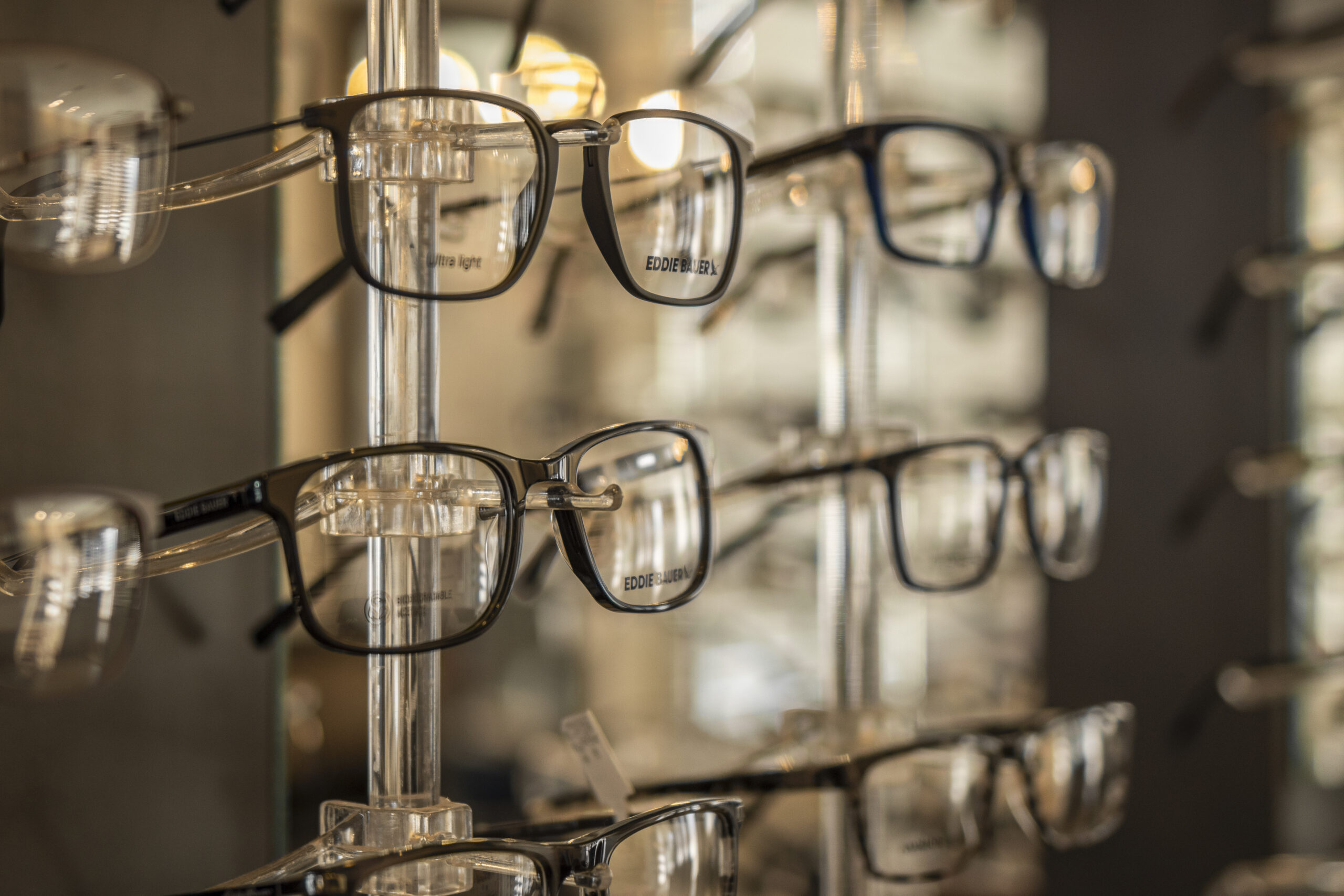 Rows of eyeglasses with black frames are displayed on a clear stand in an optical store.