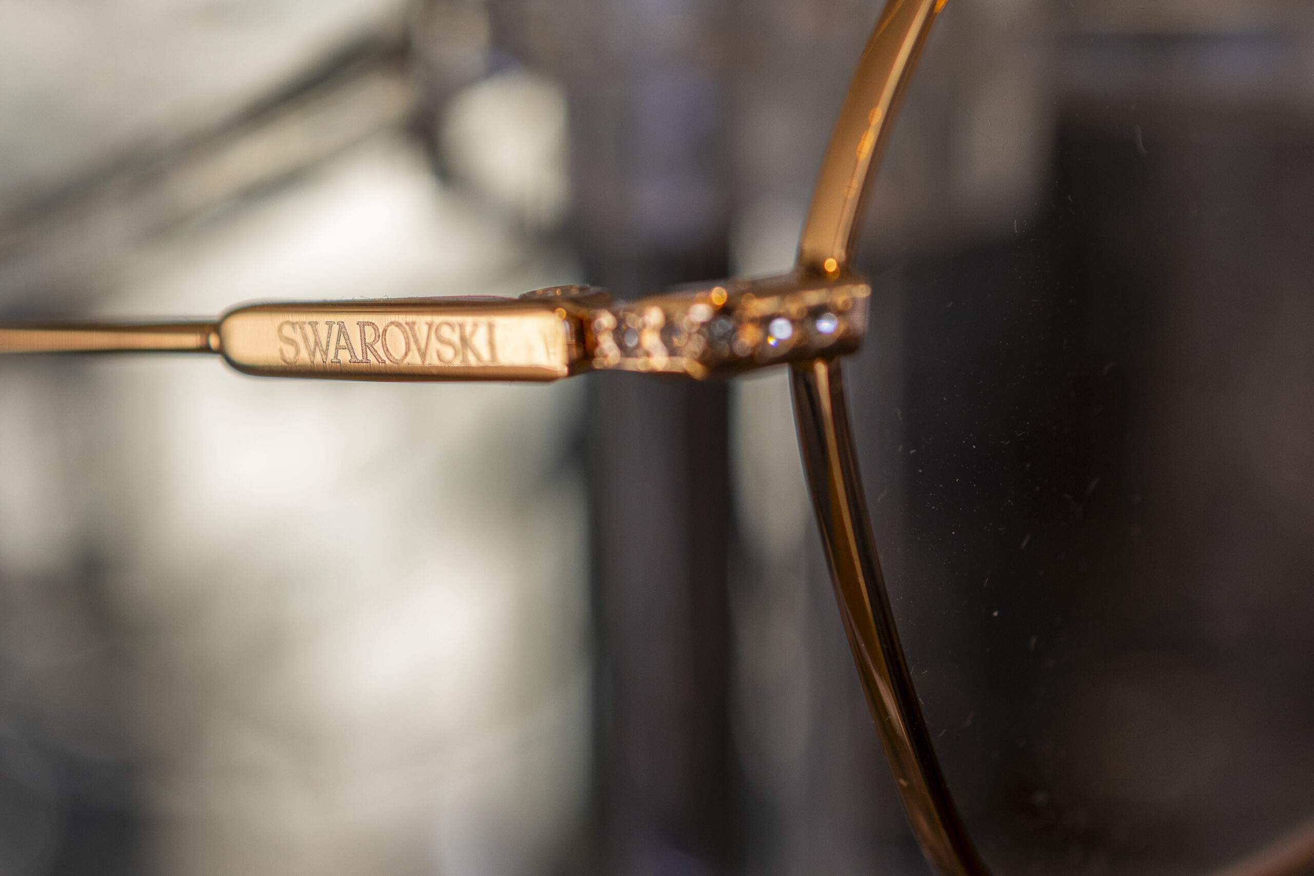 Close-up of the side arm of gold-rimmed eyeglasses with "SWAROVSKI" engraved on it, featuring small decorative crystals near the hinge.