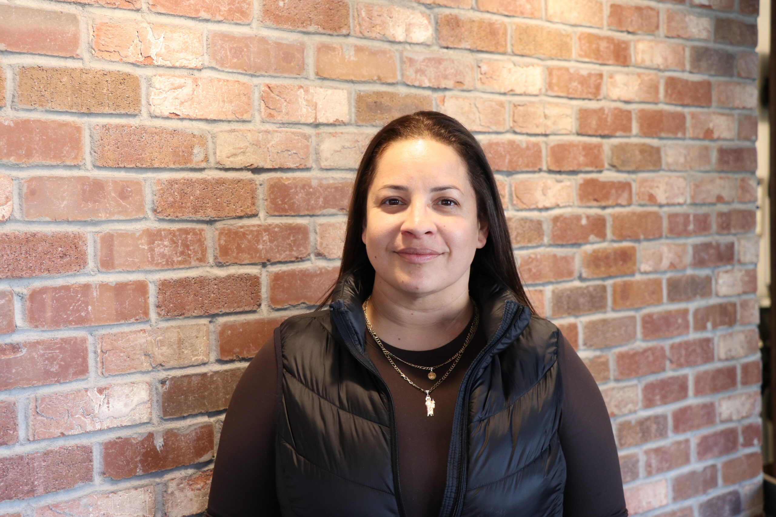 Spectrum Eye Care – Sasha A woman with long dark hair, wearing a black puffer vest over a brown shirt, stands in front of a brick wall, looking at the camera and smiling.