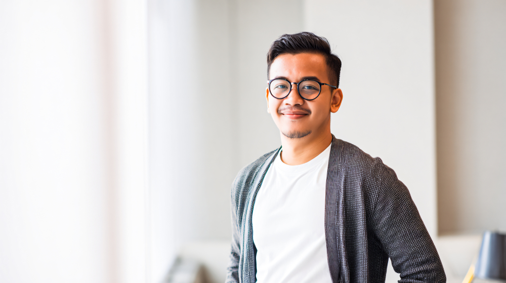 A man wearing glasses, a white t-shirt, and a gray cardigan stands indoors, smiling at the camera with a neutral background.