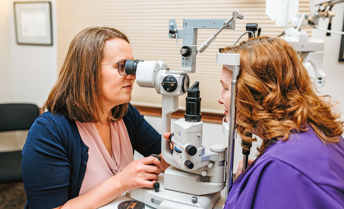 Dr. Maciejewski Spectrum Eye Care Two women sit facing each other as one conducts an eye examination using a slit lamp instrument in a clinical setting.