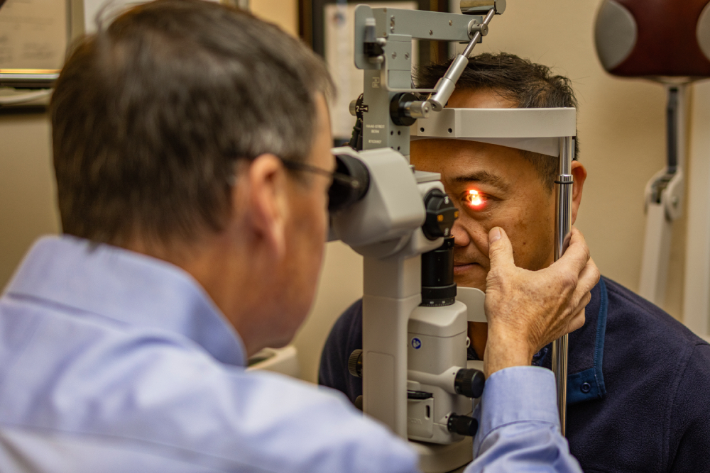 An eye doctor performs an eye exam on a patient using a slit lamp, shining a bright light into the patient's eye.