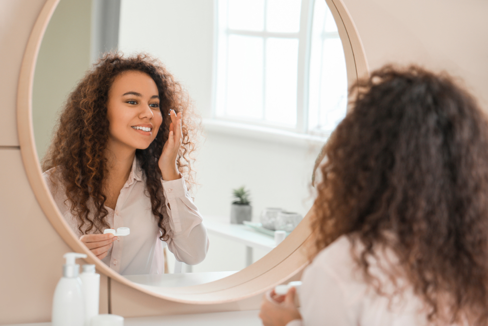 The Contact Lens Fitting Process A woman with curly hair applies cream to her face while looking in a round mirror in a bright bathroom.