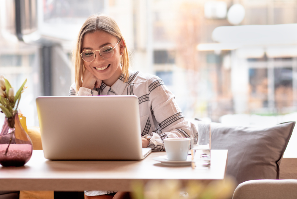 Smiling woman wearing glasses sits at a table in a cafe, looking at a laptop. There is a cup, saucer, and glass on the table.