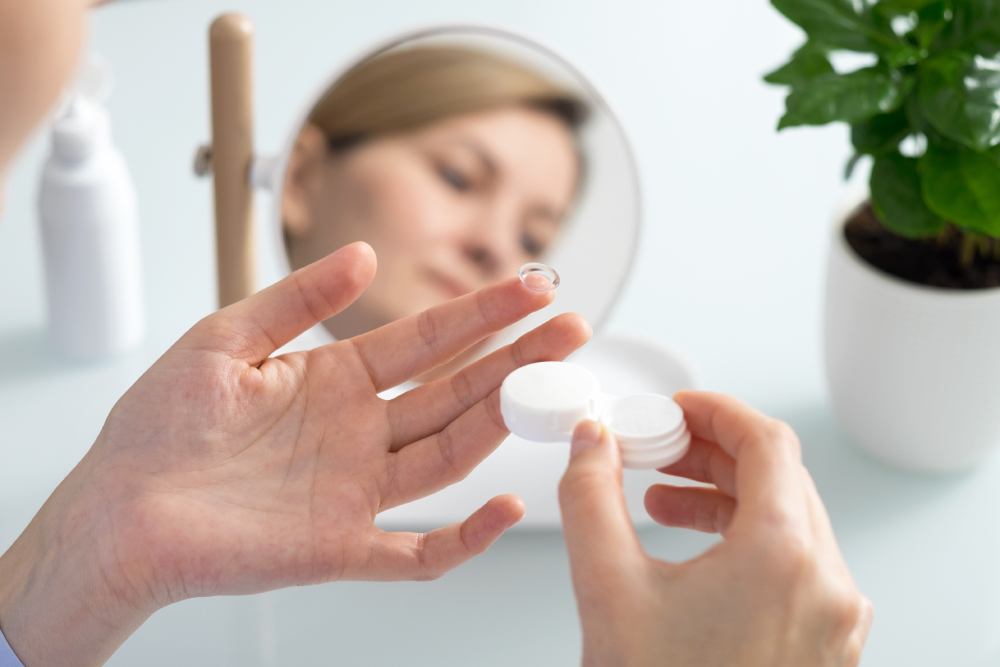 Vision Conditions Corrected With Contact Lenses A person holds a contact lens above a case with a mirror and a potted plant in the background.