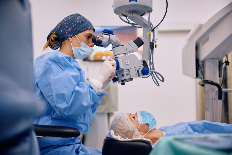 A medical professional in surgical attire uses a microscope to perform an eye procedure on a patient lying on an operating table.