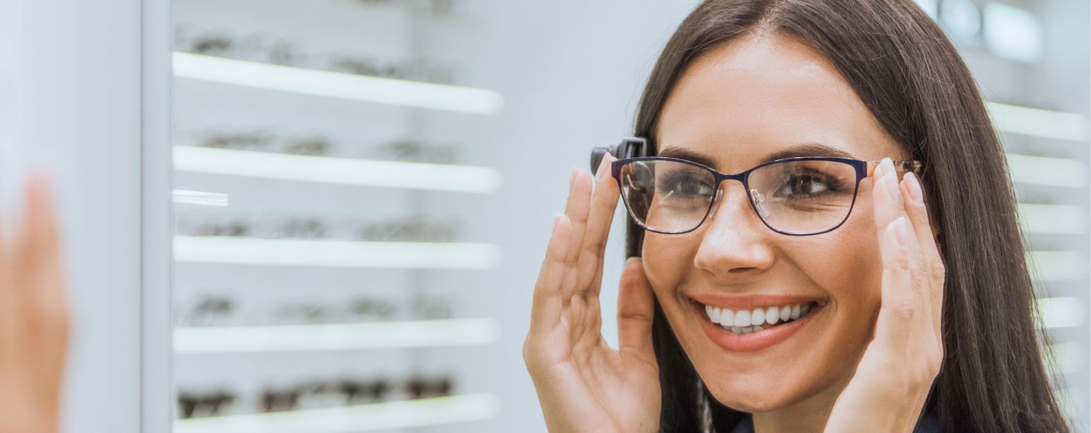 A woman smiles while trying on eyeglasses in an optical store, with display shelves of glasses visible in the background.