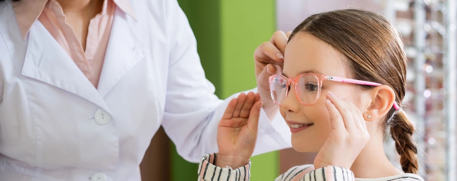An optometrist helps a young girl try on pink eyeglasses during an eye exam in an optical store.