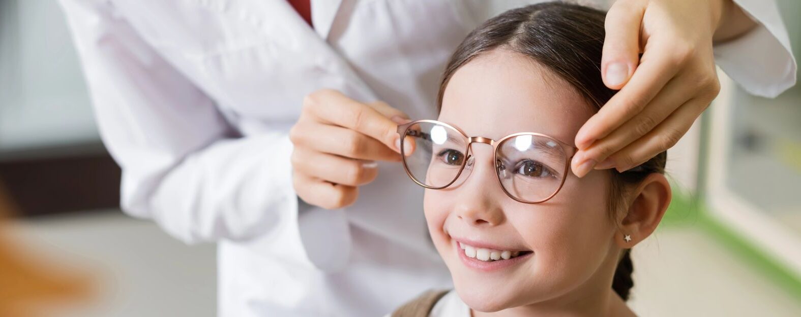 An optometrist fits eyeglasses on a smiling young girl during an eye examination.
