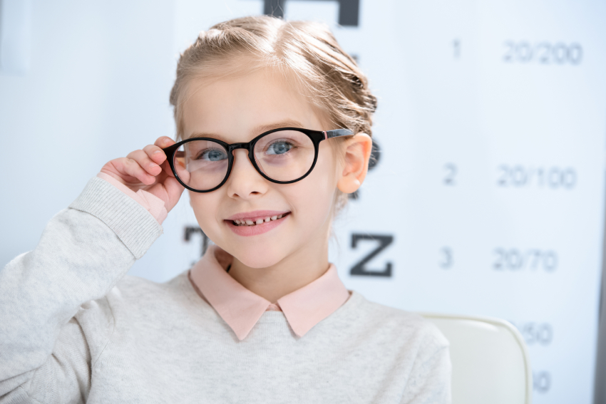Young girl wearing glasses and smiling, seated in front of an eye chart at an optometrist’s office.