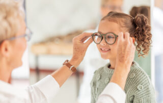 An older person helps a smiling young girl try on eyeglasses in what appears to be an optical store or clinic.