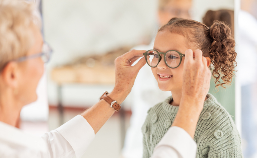 An older person helps a smiling young girl try on eyeglasses in what appears to be an optical store or clinic.