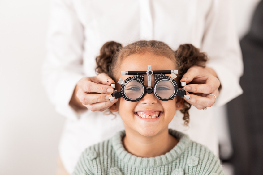 A child smiling while wearing a phoropter during an eye exam, with an adult adjusting the device from behind.