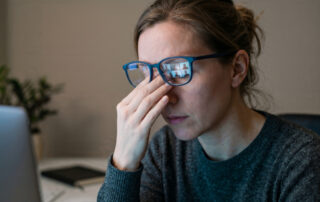 A person wearing glasses sits at a desk, rubbing their eyes while looking at a laptop, appearing tired or stressed—highlighting the importance of pediatric eye care for managing children's vision during screen time.