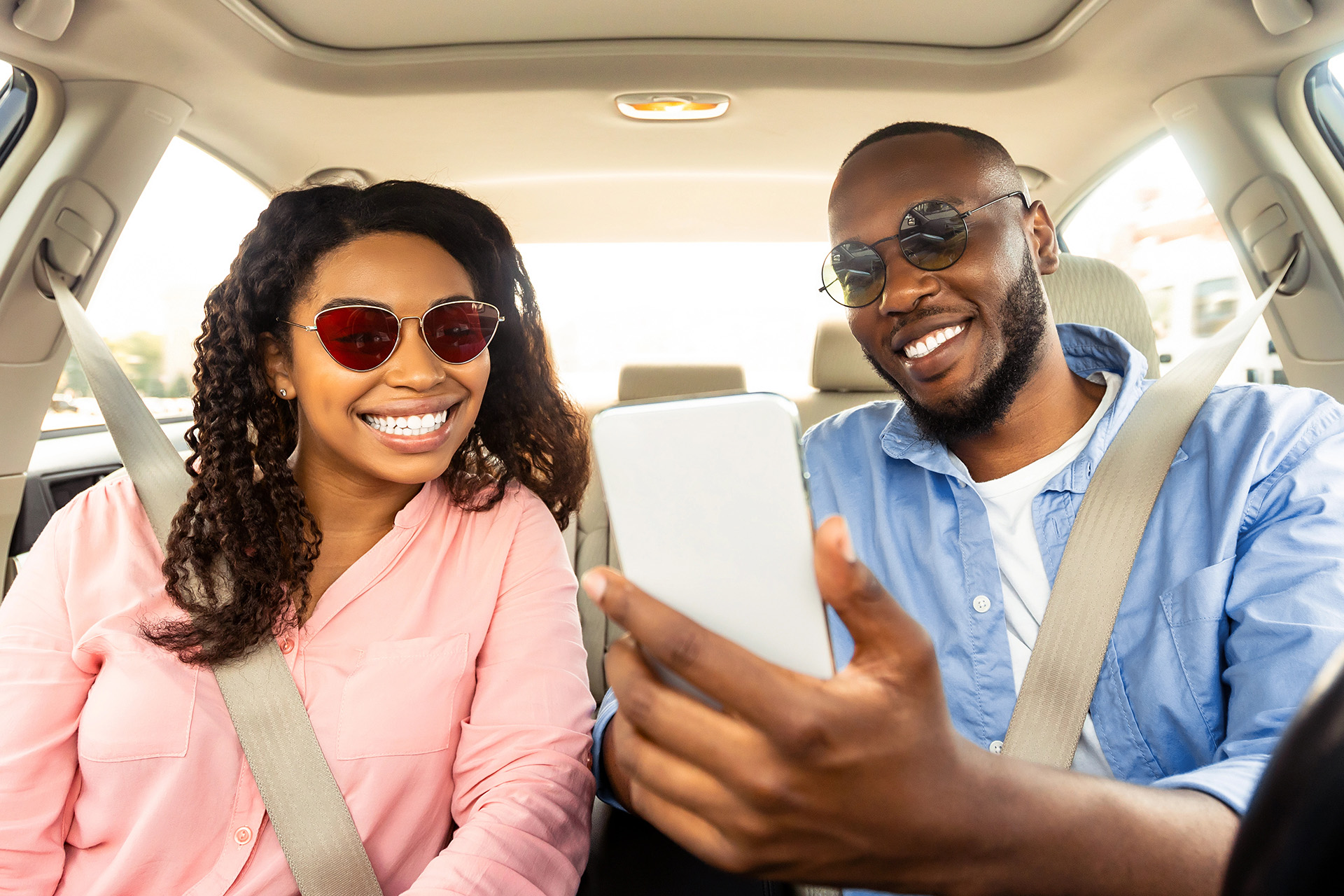 Prescription Sunglasses Car Two people sit in a car wearing seatbelts and sunglasses, smiling while looking at a smartphone held by the person in the passenger seat.
