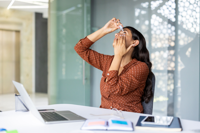 A woman sits at a desk in an office, tilting her head back and applying eye drops, with a laptop and open notebook in front of her—reminding us of the importance of regular breaks and pediatric eye care for maintaining childrens vision.