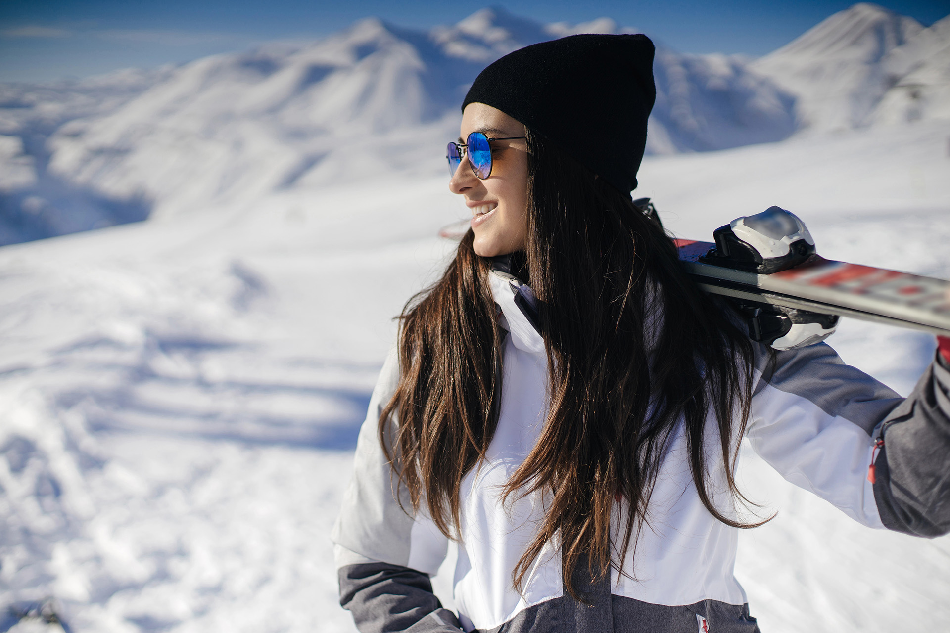 Woman skiing with prescription sunglasses A woman wearing sunglasses and a black beanie carries skis on her shoulder while standing in a snowy mountain landscape.